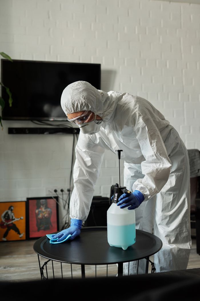 services-02 Person in protective gear cleaning a living room table with disinfectant.