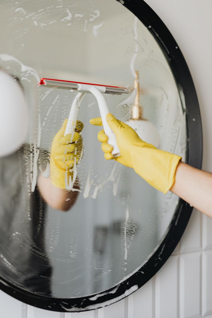 services-01 Faceless housekeeper in yellow rubber gloves cleaning round mirror from foam by squeegee in bathroom